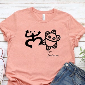 May include: A peach-colored t-shirt with a black graphic of a frog and a sun with the word "Taino" underneath.