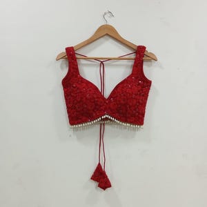 May include: A red, sleeveless top with a deep V-neck and detailed floral embroidery. The top features small white beads along the neckline and a matching red tie with decorative tassels. It is displayed on a wooden hanger.