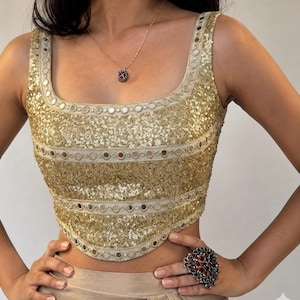 May include: A sleeveless gold crop top featuring dense sequin embellishments and horizontal bands of circular mirror work. The garment has a square neckline and a structured, fitted silhouette with a curved hemline at the waist.