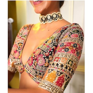 May include: A black, embroidered blouse with a deep V-neck and three-quarter sleeves. The blouse features intricate floral and geometric patterns in shades of pink, yellow, green, and gold. A black choker necklace with white pearls completes the look.