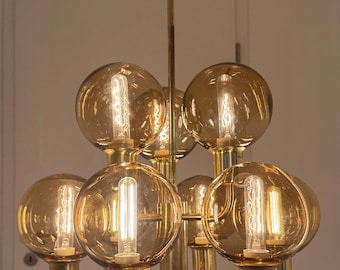 May include: A vintage brass chandelier with eight globe-shaped amber glass shades. Each shade houses a visible filament light bulb. The fixture has a central stem and curved arms, creating a mid-century modern aesthetic.