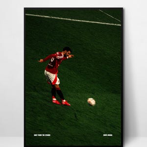 Puede incluir: Impresión enmarcada de un futbolista con uniforme rojo pateando un balón de fútbol blanco y negro en un campo verde. El jugador está en pleno lanzamiento, con el balón en movimiento. Texto "SHOT FROM THE STANDS" y "MUFC VISUAL" en la parte inferior.