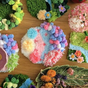 May include: A collection of colorful, handmade coasters and decorative pieces. The coasters feature various designs, including floral arrangements, landscapes, and abstract patterns, crafted with yarn and pom-poms. The color palette includes pink, blue, green, and yellow.