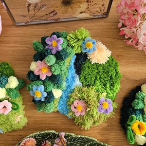 Op de afbeelding: Een verzameling kleurrijke, getextureerde bloemenkunstwerken, waaronder een krans en diverse decoratieve onderzetters. De stukken hebben verschillende kleuren, waaronder groen, blauw, roze en geel, en zijn gerangschikt op een houten oppervlak.