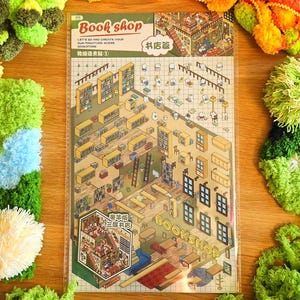 Op de afbeelding: Een kleurrijke illustratie van een boekwinkel met de tekst "Book shop" en een gedetailleerde interieurscène. Rondom de illustratie bevinden zich handgemaakte groene en gele pompom-decoraties, die een levendige en speelse uitstraling creëren.