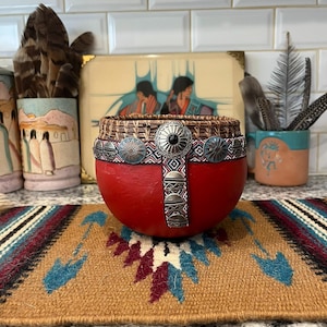 May include: A round, red gourd bowl featuring a woven rim and a decorative band adorned with silver-toned metal conchos and a central black stone. The band includes a vertical strip with geometric patterns and additional small silver metal accents.