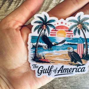 May include: A sticker featuring a coastal scene with palm trees, an eagle in flight, and another perched on the beach. A surfboard with a red, white, and blue design is also present. The text "The Gulf of America" is displayed.