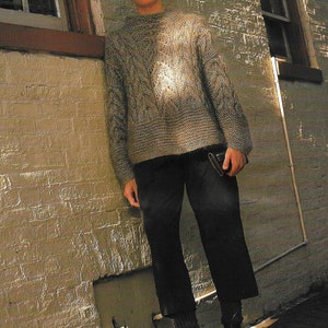 May include: A woman wearing a grey cable knit jumper with a brown leather purse in her hand. She is standing in front of a brick wall and wearing black trousers and black boots.
