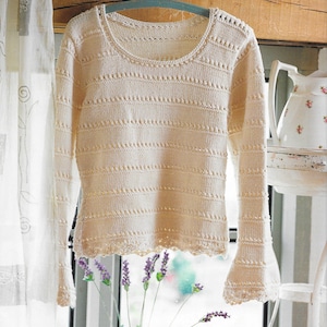 Puede incluir: Un suéter de punto blanco con un delicado patrón de encaje y un escote redondo. El suéter tiene un dobladillo y puños festoneados.