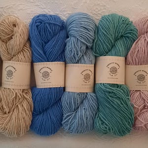 Può includere: Una collezione di otto matasse di filato in vari colori, tra cui nero, beige, blu, verde acqua, rosa e viola. Ogni matassa è avvolta con una fascia di carta con il testo "Connett Works, Fine Cut". Il filato è disposto su una superficie bianca strutturata.