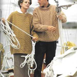May include: Two people wearing brown cable knit sweaters stand on a boat. The woman is holding a rope and the man is holding a rope and a white hat.