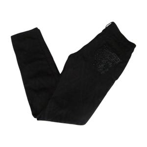 May include: Black denim pants with a decorative design on the back pocket. The pants are folded and displayed on a white background. The design on the pocket appears to be made of small, dark-colored embellishments.