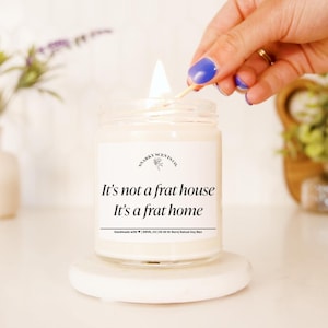 May include: A white candle with a black label that reads "It's not a frat house It's a frat home". The candle is lit and a hand is holding a match to light it.