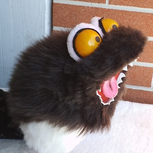 May include: A brown and white furry puppet with a large, open mouth and a pink tongue sticking out. The puppet has two large, yellow eyes with black pupils.