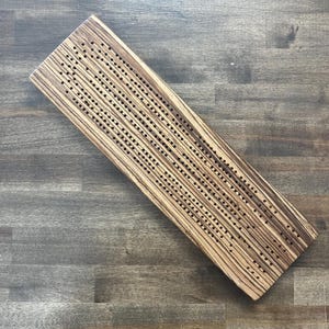 May include: A wooden cribbage board with a striped wood grain pattern. The board has holes for pegs and is ideal for playing the game of cribbage.