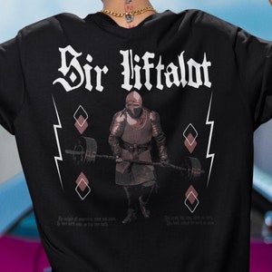 Puede incluir: Camiseta negra con un gráfico de un caballero levantando una barra. El texto "Sir Liftalot" está impreso en la camiseta.