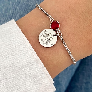 May include: A silver bracelet featuring a round charm with an engraved family illustration and a red gemstone. The bracelet is worn on a wrist, with a white sleeve and blue denim in the background.