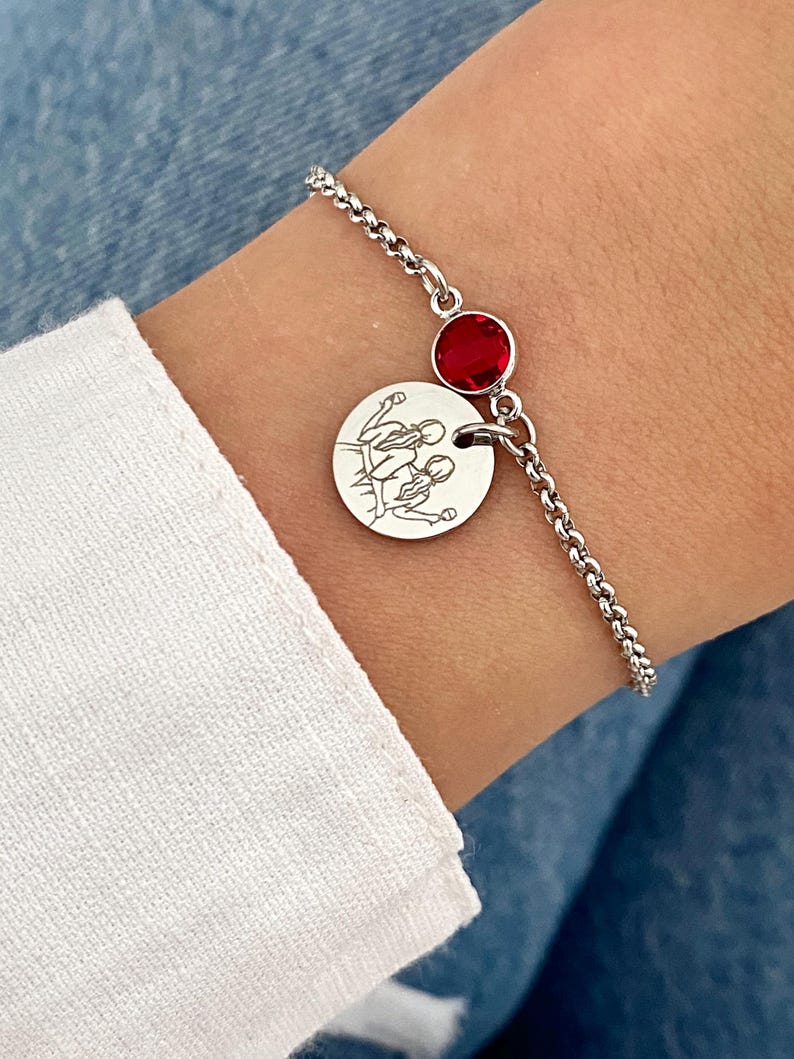 May include: A silver bracelet featuring a round charm with an engraved family illustration and a red gemstone. The bracelet is worn on a wrist, with a white sleeve and blue denim in the background.