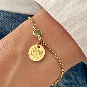 May include: Gold-colored bracelet featuring a small green gemstone and a round charm with a hand-drawn illustration of two hands interlocked. The bracelet is worn on a wrist, with a white shirt and denim jeans in the background.