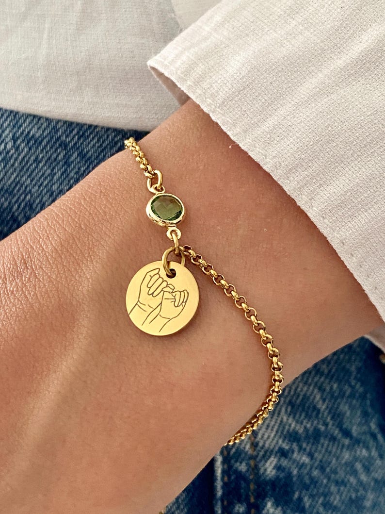 May include: Gold-colored bracelet featuring a small green gemstone and a round charm with a hand-drawn illustration of two hands interlocked. The bracelet is worn on a wrist, with a white shirt and denim jeans in the background.