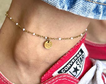 May include: Gold-colored anklet with small white beads and a circular charm featuring a heart design. The anklet is worn on an ankle, paired with red sneakers and blue denim jeans.