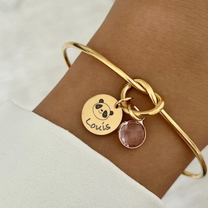 May include: Gold bangle bracelet with a knot design and a charm featuring a panda face and the name "Louis". The charm is attached to a pink crystal.