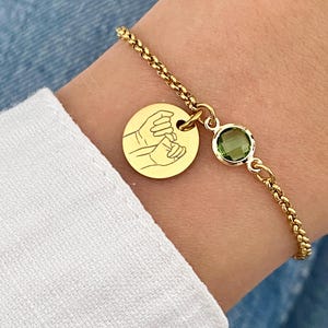 May include: Gold-toned bracelet featuring a round charm with a hand-holding design and a green gemstone. The bracelet has a delicate chain and is worn on a wrist with a white sleeve.