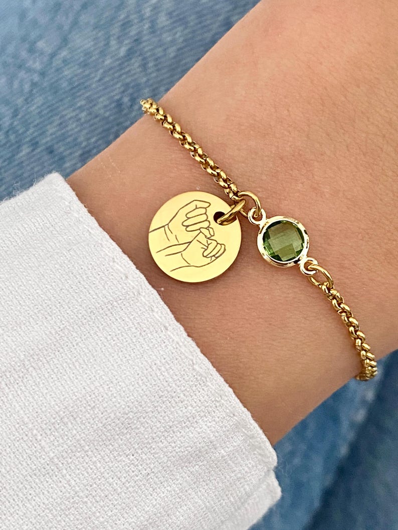 May include: Gold-toned bracelet featuring a round charm with a hand-holding design and a green gemstone. The bracelet has a delicate chain and is worn on a wrist with a white sleeve.