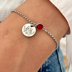 May include: A silver bracelet with a round charm featuring a line drawing of three figures. A small, round, red gemstone is set near the clasp. The bracelet is worn on a wrist, with a white shirt and blue jeans in the background.