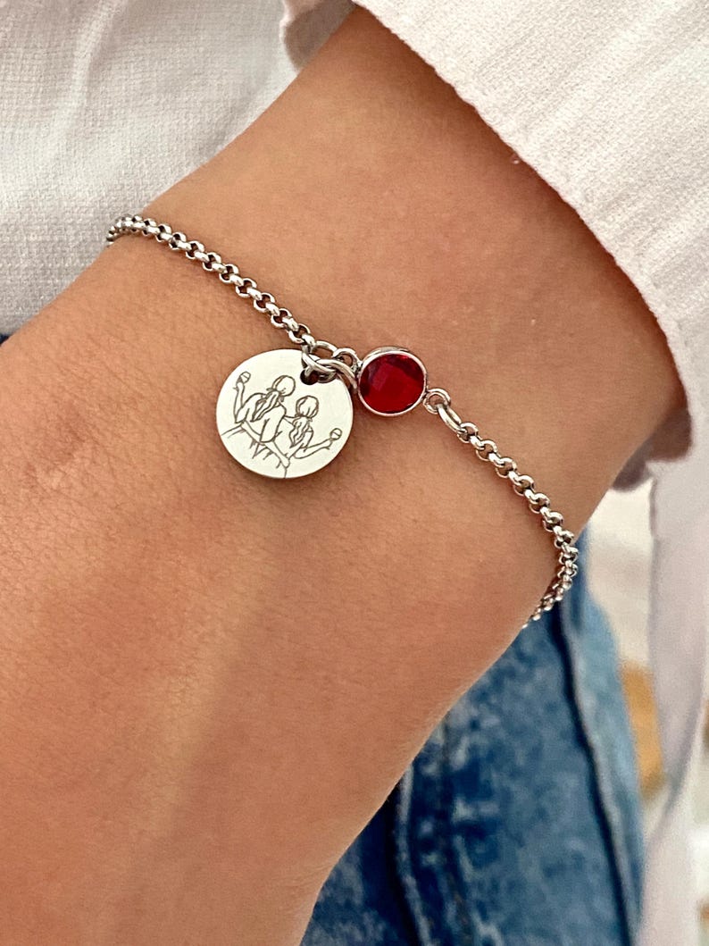 May include: A silver bracelet with a round charm featuring a line drawing of three figures. A small, round, red gemstone is set near the clasp. The bracelet is worn on a wrist, with a white shirt and blue jeans in the background.
