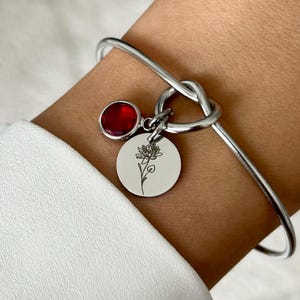 Puede incluir: Un brazalete de plata con un diseño de nudo y un dije con una piedra roja y una flor grabada delicada.