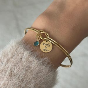 May include: A gold-toned bangle bracelet featuring a central knot design. Attached to the knot are two charms: a small, round, teal-colored stone and a circular gold-toned disc engraved with the text "Emily & Alice" in a black script font.