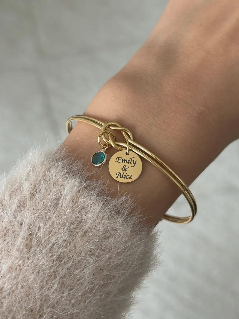 May include: A gold-toned bangle bracelet featuring a central knot design. Attached to the knot are two charms: a small, round, teal-colored stone and a circular gold-toned disc engraved with the text "Emily & Alice" in a black script font.