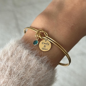 May include: Gold bangle bracelet with a knot charm and a small blue gemstone. The charm has the inscription "Emily & Alice".