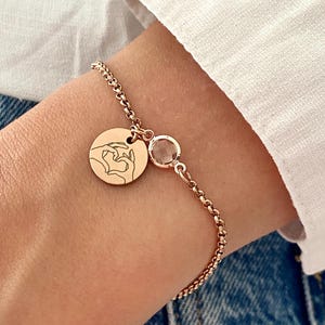 May include: Rose gold bracelet featuring a round charm with a hand-heart design and a small, round, clear gemstone. The bracelet is worn on a wrist, with a white shirt and denim jeans in the background.