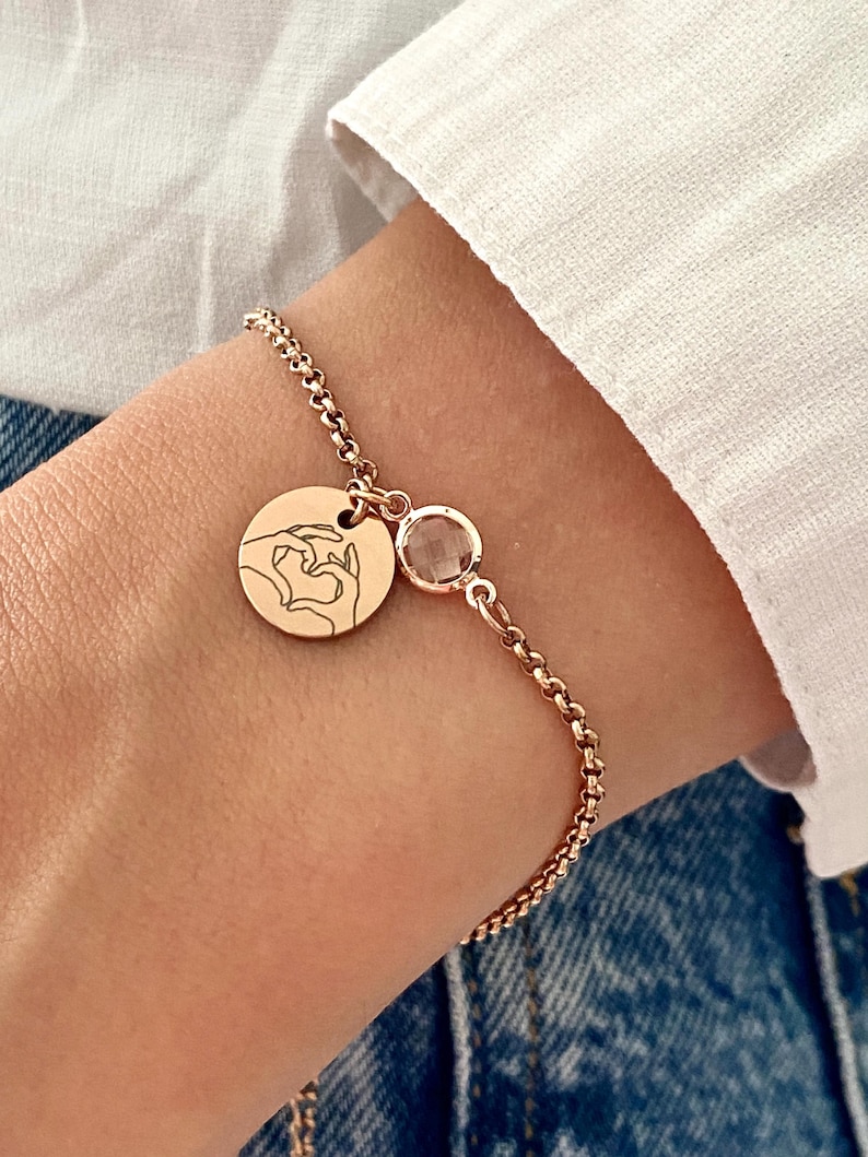 May include: Rose gold bracelet featuring a round charm with a hand-heart design and a small, round, clear gemstone. The bracelet is worn on a wrist, with a white shirt and denim jeans in the background.