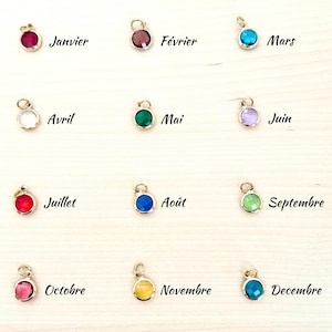 May include: A collection of twelve gold-colored pendants, each featuring a different colored gemstone. Each pendant has a small loop at the top and is labeled with a month of the year in French. The gemstones are various colors, including red, blue, and green.