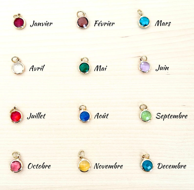 May include: A collection of twelve gold-colored pendants, each featuring a different colored gemstone. Each pendant has a small loop at the top and is labeled with a month of the year in French. The gemstones are various colors, including red, blue, and green.