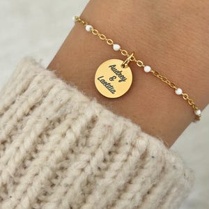Pode incluir: Um bracelete de corrente dourada com contas brancas e um pingente redondo gravado com "Audrey & Laetitia".