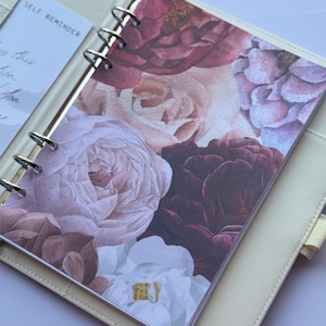 Puede incluir: Agenda de color crema con un inserto floral con rosas de acuarela en tonos burdeos, rosa y blanco. La agenda está abierta, revelando una tarjeta "Self Reminder". Los anillos de encuadernación son plateados.
