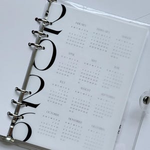 May include: A clear, plastic binder with a 2026 calendar insert. The calendar pages are white with black text, displaying the months and dates. The binder has silver metal rings and a snap closure.