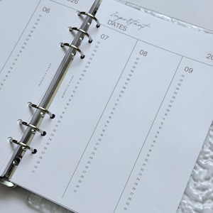 Op de afbeelding: Een close-up van een open planner met een doorzichtige kaft. De pagina's zijn wit met grijze lijnen en de tekst "Important Dates" bovenaan. De planner is open op een pagina met de maand februari 2026 en genummerde dagen.