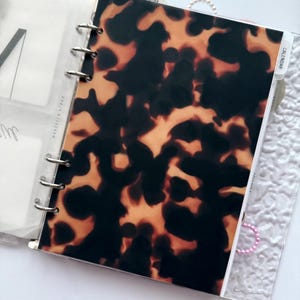 May include: A planner with a tortoiseshell pattern cover. The cover is brown and black with a glossy finish. The planner is open, revealing a calendar tab and other pages. The planner is held together with silver rings.