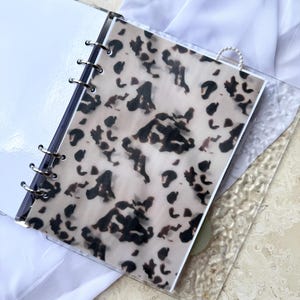 May include: A clear binder with silver rings, holding a sheet with a tan and black animal print design. The binder is open, revealing white pages. A clear, textured sheet is visible on the right side.
