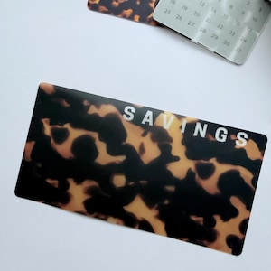 May include: A card with a tortoiseshell design and the word "SAVINGS" in white. A 2026 calendar is in the background, along with a card with the same pattern. The card is rectangular.