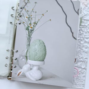 Peut inclure: Un agenda sur le thème de Pâques. L'image présente un œuf vert clair avec des détails de feuilles, posé sur un support blanc en forme de lapin. Un vase de fleurs blanches et un calendrier sont également visibles. L'agenda est ouvert, révélant une page transparente.