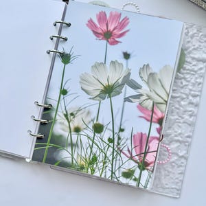 May include: A planner with a floral insert featuring pink and white cosmos flowers against a blue sky. The planner has a clear cover and silver metal rings. A calendar is visible in the background.