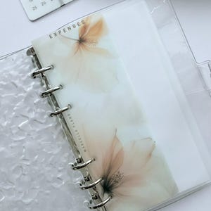 May include: Clear, textured planner with a floral design on the "Expenses" divider. The planner has silver metal rings and a calendar in the background. The floral design is in shades of beige and peach.