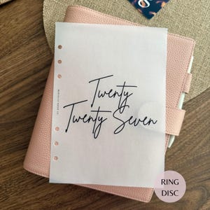 May include: A translucent vellum sheet with the black text Twenty Twenty Seven printed in a script font, featuring a vertical row of six circular punch holes along the left edge, resting atop a textured light pink leather ring binder planner.