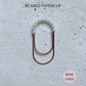May include: A rose gold metal paperclip with a U-shaped design, featuring a row of small white beads along the top curve. The text BEADED PAPERCLIP is printed at the top, and the text ROSE GOLD is printed in a circle at the bottom right corner.